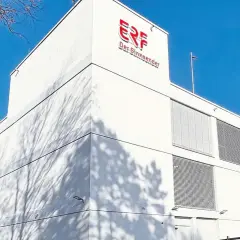Das Gebäude von ERF, dem Sinn-Sender, unter blauem Himmel. Schatten von Bäumen tanzen auf der weißen Fassade.