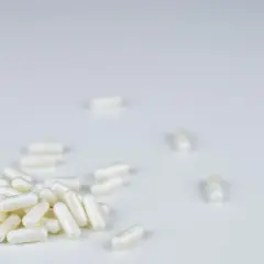 Garść białych kapsułek, jak anapran, rozsypana na białym tle. Kilka pojedynczych kapsułek leży dalej.