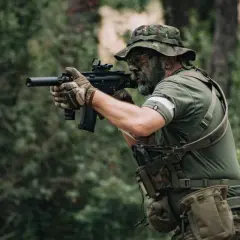 Jugador de deporte airsoft con camuflaje facial y equipo táctico, apuntando con su réplica de rifle en un bosque.
