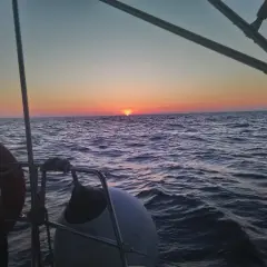 Zachód słońca nad wzburzonym oceanem atlantyckim widziany z pokładu żaglówki. Widać koło ratunkowe i ster.
