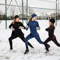 Trzy dziewczynki w czarnych i granatowych koszulkach termoaktywnych dziecięcych ćwiczą na śniegu przy boisku.