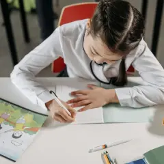Dziewczynka pisze w zeszycie, obok leży pluszowy piesek i książeczka z rysunkami dzieci puszczających latawce. To jak nauka a2 - b2.