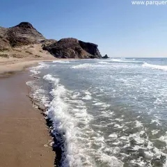 Olas rompiendo en la arena dorada de la playa del Barronal, con montañas áridas al fondo bajo un cielo azul.