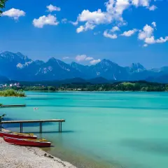 Türkisblaues Wasser am Badesee Bayern, mit roten Ruderbooten am Kiesstrand und majestätischen Alpen im Hintergrund.