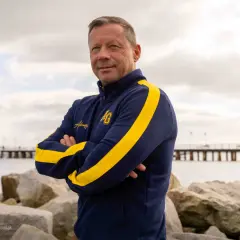 Mężczyzna w granatowej bluzie z żółtymi pasami, z rękami skrzyżowanymi na piersi. To trener Arki Gdynia, pozujący na tle morza i molo.