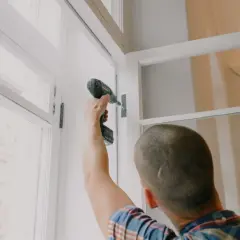 Mężczyzna przykręca zawias w nowym oknie. Planujesz wymiana okien? Cennik usług montażowych znajdziesz w internecie.
