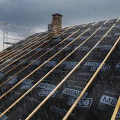 Regen prasselt auf ein Dach, dessen Unterspannbahn von innen repariert wird. Holzlatten sind auf der schwarzen Folie befestigt.