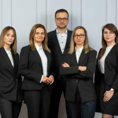 Pięć osób w garniturach, zespół prawników, stoi przed jasną ścianą.