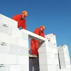Dwóch budowlańców w pomarańczowych strojach montuje betonowy nadproże nad otworem w ścianie z białych pustaków.