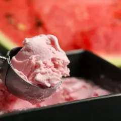 Un cucchiaio raccoglie una pallina di cremoso gelato anguria fatto in casa, con una fetta di anguria sullo sfondo.