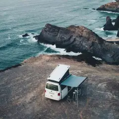 Furgoneta camper con toldo desplegado en un acantilado, disfrutando del camping en el norte de Portugal. El mar azul choca contra las rocas.