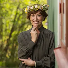 En kvinna med blomsterkrans ler mot kameran. Hon påminner om en sommarbild av mischa billing familj.
