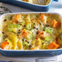 Herzhaft überbackener Lachs mit Kartoffeln in einer blauen Auflaufform, garniert mit Frühlingszwiebeln und rosa Pfefferbeeren.
