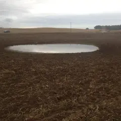 Pola po orce, kałuża i pracujący traktor. To obrazek pokazujący, melioracja co to, czyli poprawę stosunków wodnych na ziemi.
