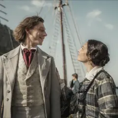 Chopin! Obsada. Mężczyzna w jasnym fraku i kobieta w kratowanej sukni patrzą na siebie na tle portu.