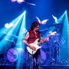 Mężczyzna gra na gitarze elektrycznej na scenie, w tle perkusja i niebieskie światła z motylami.