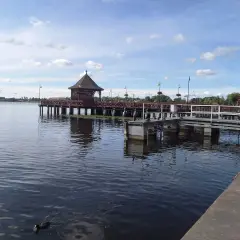 Drewniana altana na molo w Ostródzie, otoczona wodą i zielenią. Kaczki pływają spokojnie.