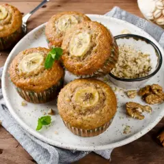 Pięć pysznych muffinek bananowych z plasterkiem banana na wierzchu, udekorowane miętą. Obok miseczka z orzechami i laska cynamonu.