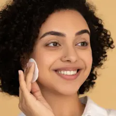 Donna sorridente con capelli ricci usa un dischetto per struccarsi, mostrando come si usa il burro struccante per una pelle luminosa.