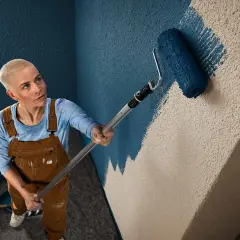 Frau mit Kurzhaarschnitt streicht mit blauem Roller eine Wand mit Rauputz.