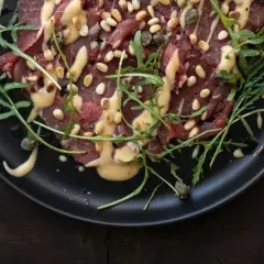 Rinder Carpaccio mit Pinienkernen, Kapern und Rucola auf schwarzer Platte, beträufelt mit einer cremigen Sauce.
