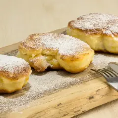 Trzy puszyste placki z jabłkami, posypane cukrem pudrem, na drewnianej desce. Idealne na najprostszy przepis na placki z jabłkami.