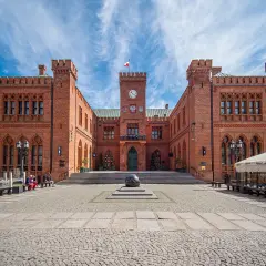 Rynek Kołobrzeg: ceglany ratusz z zegarem i flagą, przed nim kamienna rzeźba i brukowany plac.
