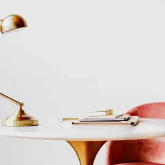 Elegante lampara de mesa metal dorada sobre un escritorio blanco con libros y una silla rosa.