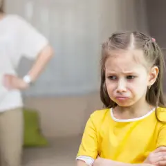 Smutna dziewczynka w żółtej bluzce, z matką w tle.