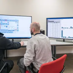 Dwóch mężczyzn analizuje dane na monitorach. Na jednym schemat budynku, na drugim wykresy, które pomagają w prognozowaniu popytu.