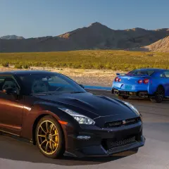 Dwa Nissan GTR R35, jeden brązowy, drugi niebieski, stoją na torze wyścigowym na tle pustynnego krajobrazu.