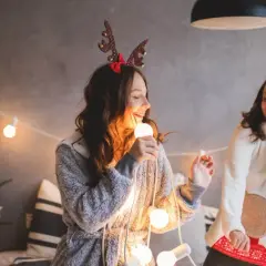 Due ragazze cantano canzoni di Natale in inglese con testo, tenendo una lampadina come microfono, circondate da luci natalizie e un albero addobbato.