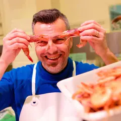 Hombre sonriente jugando con mariscos rojos, sosteniendo dos gambas frente a sus ojos.