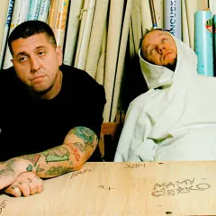 Dwóch mężczyzn siedzi przy drewnianym stole. Jeden w czarnej koszulce, drugi w białej bluzie z kapturem.