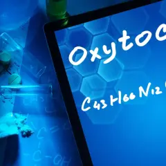 Na ekranie tabletu widnieje napis "Oksytocyna co to" i jej wzór chemiczny. W tle leżą tabletki, proszek i probówki.