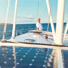 Un uomo sorride sul ponte di una barca a vela, con un impianto pannello solare che riflette il cielo azzurro.