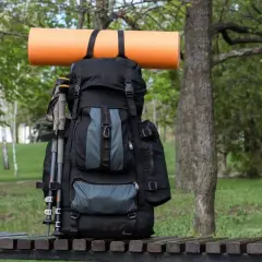 Ein schwarzer Rucksack mit Wanderstöcken und einer orangefarbenen Isomatte, die mit Schlaufen befestigt ist, steht auf einer Holzbank im Grünen.