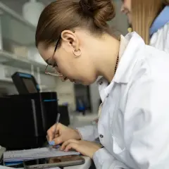 Studentka w białym fartuchu laboratoryjnym pisze notatki w zeszycie, obok klawiatury i telefonu.