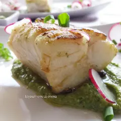 Un trozo de pescado blanco y tierno reposa sobre una cama de vibrante mojo verde, adornado con rodajas de rábano y cebolleta.