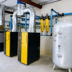 Instalación industrial con compresores y secadores de aire comprimido, tuberías azules y un gran tanque de almacenamiento plateado.