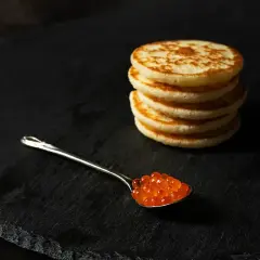 Une cuillère de caviar rouge sur une assiette noire, à côté d'une pile de blinis dorés. Parfait pour une recette blinis gourmande.
