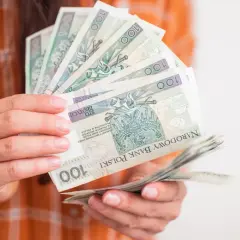 Dłonie trzymają wachlarz banknotów 100 zł. Taka kwota to już całkiem spora kuroniówka na drobne wydatki.