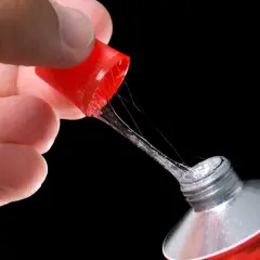 Main d'une personne tenant le bouchon rouge d'un tube de colle forte, avec des fils de colle s'étirant. Astuce pour comment enlever de la colle forte sur du métal.