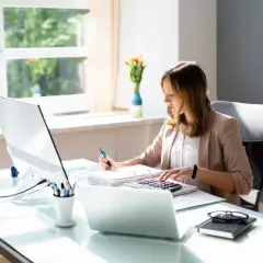 Kobieta pracuje przy biurku, korzystając z kalkulatora i notując. Jej praca jest zgodna z polityką rachunkowości firmy.