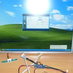 Na biurku leży monitor z pulpitem Windows XP, obok kable i płytka sterująca. To może być pomocne, jak podłączyć monitor do laptopa.