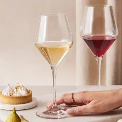 Dwie lampki wina, białe i czerwone, obok tartaletki z bezą i owoców. Dłoń z pierścionkiem zbliża się do kieliszka.