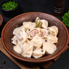 Pielmieni co to? Pyszne, domowe pierożki z mięsem, podane w glinianej misie z liśćmi laurowymi i pieprzem. Obok śmietana i natka pietruszki.