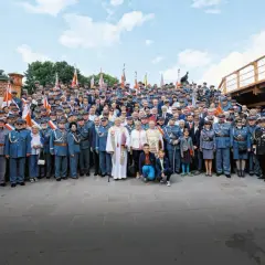 Grupa osób w historycznych mundurach, przypominających przedwojenne stopnie wojskowe, pozuje do zdjęcia na tle ceglanej budowli i drewnianej konstrukcji.