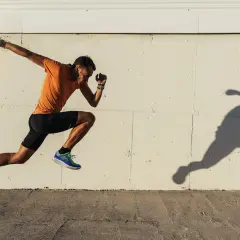 Mężczyzna w pomarańczowej koszulce i czarnych spodenkach wykonuje trening szybkościowy, unosząc się w powietrzu. Jego cień odbija się na jasnej ścianie.
