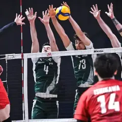 Zacięta akcja siatkarska: polska Japonia w akcji. Zawodnik w czerwonej koszulce atakuje, blokujący w zielonych strojach próbują zatrzymać piłkę.